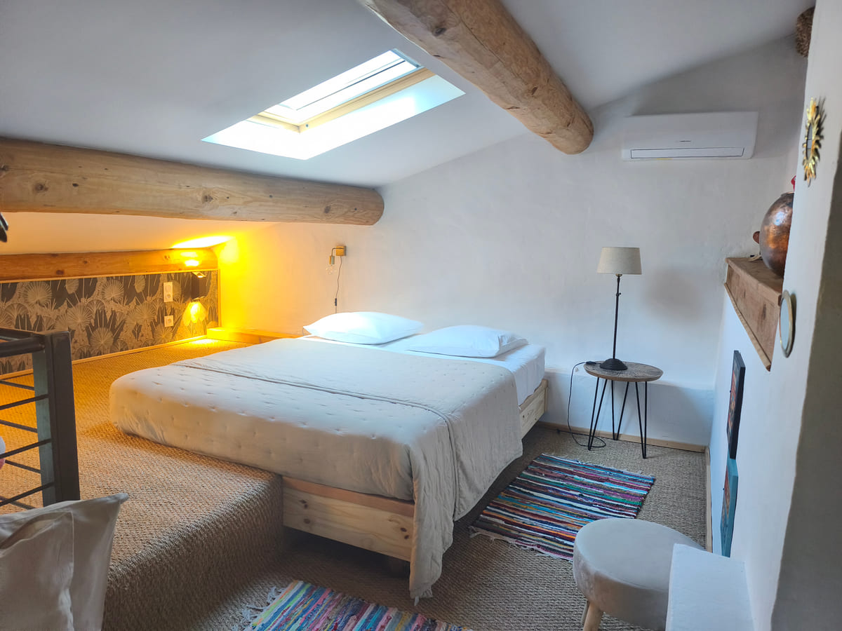 Chambre en mezzanine : lit double, poutres en bois brut et velux plein ciel